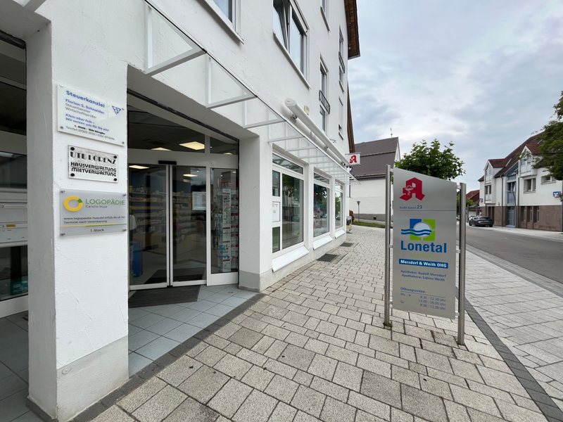 Außenansicht der Lonetal Apotheke mit Eingangstür, Apothekenschild und Aufsteller mit Öffnungszeiten und Apothekenlogo. Rechts daneben verläuft eine g